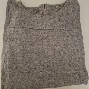 Grå långärmad stickad tröja - Mysig grå stickad tröja med lång ärm och rund hals. Tröjan har en snygg söm över bröstet och ribbade partier på ärmarna samt en dragkedja i nacken. Perfekt för lager-på-lager och chill dagar.