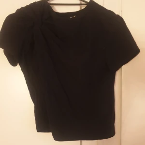 Svart topp med knut från Divided - Svart kortärmad topp från Divided i mjuk bomull. Toppen har en cool knutdetalj på ena axeln som ger en unik twist till den annars enkla designen. Perfekt för dig som gillar stilrena plagg med en liten extra detalj.