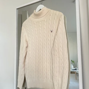Beige kabelstickad polotröja GANT - Kabelstickad polotröja från GANT i en snygg beige färg. Tröjan har hög krage, lång ärm och klassisk GANT-logga broderad på bröstet. Perfekt för dig som gillar en stilren och tidlös look med lite preppy känsla.