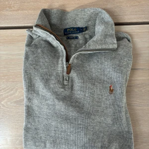 Grå half zip tröja Polo Ralph Lauren - Snygg grå långärmad tröja från Polo Ralph Lauren med broderad logga på bröstet. Tröjan har half zip med läderdetalj på dragkedjan och är ribbstickad för en klassisk look. Perfekt för lager-på-lager under höst och vinter.