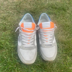 Philippe Model sneakers i vitt, grått, orange & blått - Snygga Philippe Model sneakers i vitt och grått med detaljer i blått och orange. Ovandel i mocka och textil, klassisk snörning och platt sula. Perfekta för dig som gillar stilrena men ändå färgstarka sneakers. Samt ungefär 3-4 cm hög sula så du blir lite längre 😉