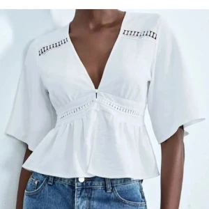 Vit blus med v-ringning och spetsdetaljer från Zara - Superfin vit blus med djup v-ringning och vida, korta ärmar. Blusen har spetsdetaljer vid byst och midja samt en lätt volang nertill. Perfekt till jeans eller kjol för en fräsch look. Luftigt material som passar till varma dagar.