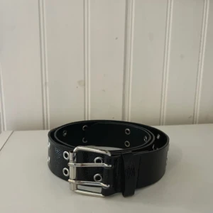 Svart bälte med nitar och dubbla spännen - Snyggt svart bälte i skinnimitation med silvriga metalldetaljer och dubbla spännen. Bältet har flera rader med metallöljetter längs hela bandet för en edgy look. Perfekt accessoar för att lyfta din outfit med lite extra attityd.