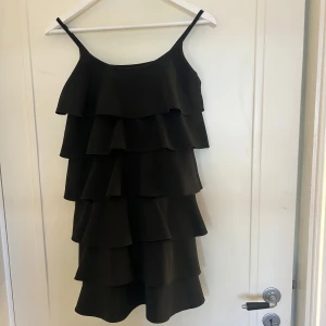Svart volangklänning med smala axelband - Supersnygg svart kortklänning med flera lager volanger och smala axelband. Perfekt för dig som vill sticka ut med en lekfull och trendig look. 