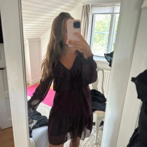 Vinröd prickig volangklänning - Vinröd kortklänning med prickigt mönster, v-ringning och volangdetaljer vid halsen. Långa ärmar i transparent tyg och resår i midjan för en skön passform. Perfekt för dig som gillar romantisk och trendig stil.