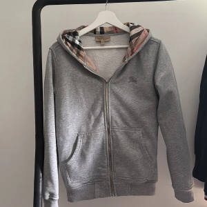 Grå hoodie från Burberry med dragkedja - Snygg grå hoodie från Burberry med klassiskt rutigt foder i huvan. Tröjan har dragkedja framtill, två fickor och långa ärmar. Perfekt för dig som gillar stilrena plagg med en touch av lyx. Ikonisk Burberry-logga broderad på bröstet. Dragkedjan fungerar halvt 