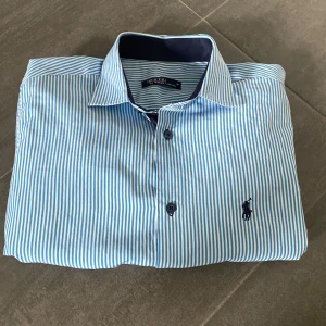 Randig skjorta från Polo Ralph Lauren - Snygg skjorta från Polo Ralph Lauren i vitt och ljusblått med smala ränder. Klassisk krage, långa ärmar och mörkblå broderad logga på bröstet. Skjortan har knäppning framtill och är tillverkad i bomull för en fräsch och stilren look.