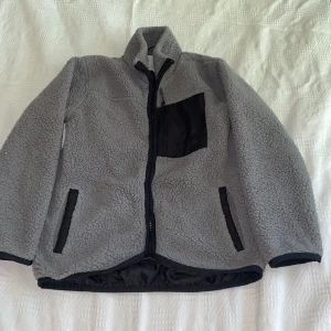 Grå teddyjacka från 157, stl 130 - Mysig grå teddyjacka från 157 med svarta detaljer och dragkedja framtill. Jackan har en svart bröstficka med dragkedja och två svarta fickor på sidorna. Perfekt för kyliga dagar och riktigt skön att ha på sig.