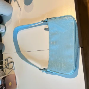 Ljusblå axelväska från Gina Tricot - Säljer en ljusblå axelväska från Gina Tricot med croco-mönstrad yta i glansigt syntetmaterial. Väskan har en stilren, rektangulär form och stängs med dragkedja upptill. Perfekt storlek för det viktigaste och en snygg detalj till din outfit.