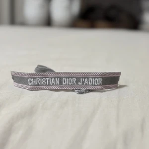 CD armband  - Superfint Cristian D armband med texten J'Adior broderat i vitt. Armbandet är grått med ljusrosa detaljer och har justerbar knytning med tofsar i ändarna. Perfekt accessoar för att lyfta din outfit med en lyxig touch. Pris kan diskuteras vid snabb affär.