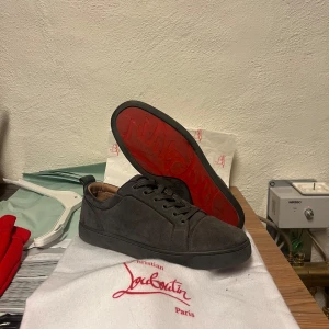 Grå mockasneakers från Louboutin - Snygga grå sneakers från Christian Louboutin i mocka med klassisk röd sula. Skorna har rund tå, platt sula och snörning. Perfekta för dig som vill ha en stilren och lyxig look med en ikonisk touch.
