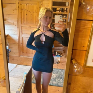 Svart kort klänning med cut-outs - Snygg svart kort klänning med långa ärmar och trendiga cut-outs framtill. Klänningen har smala axelband som korsas över bröstet och ryggen, vilket ger en cool och modern look. Tight passform som framhäver figuren, perfekt för dig som gillar att sticka ut.