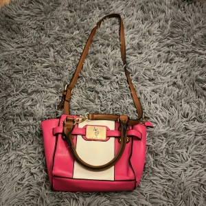 Rosa och vit axelväska från U.S. Polo Assn. - Säljer en snygg axelväska från U.S. Polo Assn. i rosa och vitt med bruna detaljer. Väskan har både handtag och justerbar axelrem, flera fack med dragkedja och guldfärgad logga framtill. Perfekt för dig som vill sticka ut med färg och stil.