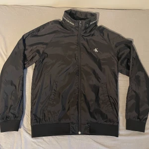 Svart vindjacka från Calvin Klein Jeans - Svart vindjacka från Calvin Klein Jeans med hög krage och logga på bröstet. Jackan har dragkedja, ribbade muddar och meshfoder på insidan. Snygg detalj med Calvin Klein Jeans-tryck runt kragen. Perfekt för dig som gillar stilren och sportig look.