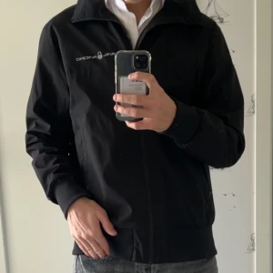 Svart vindjacka från Sail Racing - Svart vindjacka från Sail Racing med dragkedja framtill och diskret logga på bröstet. Jackan har hög krage, långa ärmar och ribbade muddar. Perfekt för dig som gillar stilren och sportig look.
