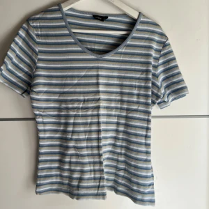 Randig t-shirt från Lindex - Superfin t-shirt från Lindex med blå, vita och beige horisontella ränder. T-shirten har korta ärmar, v-ringning och är gjord i mjuk bomull. Perfekt för en chill och stilren look.