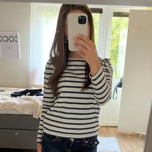 Randig långärmad tröja med dragkedja - Säljer en vit och marinblå randig långärmad tröja med snygga puffade axlar och dragkedja bak i nacken. Tröjan har en klassisk båtringad hals och är gjord i mjuk bomull, perfekt för en chill och stilren look.