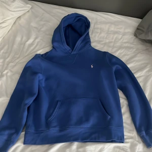Blå hoodie från Polo Ralph Lauren - Snygg blå hoodie från Polo Ralph Lauren med klassisk broderad logga på bröstet. Tröjan har huva, känguruficka och långa ärmar. Perfekt för dig som gillar stilrena och bekväma plagg. Materialet är mjuk bomull som känns skönt mot huden.