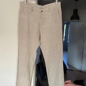Beige linnebyxor från NN07 - Snygga beige linnebyxor från NN07 med klassisk passform och raka ben. Byxorna har bälteshällor, sidofickor och två bakfickor. Perfekta för en chill och stilren look under varma dagar. Fint sydda där de tidigare var hål.