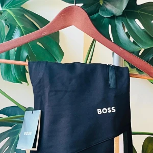 Svarta shorts från BOSS med rand - Svarta shorts från BOSS med vit logga på ena benet och en snygg rand i vitt och gult längs sidan. Modellen har dragkedja upptill och sportig känsla. Perfekta för dig som gillar stilrena och trendiga plagg med en twist.