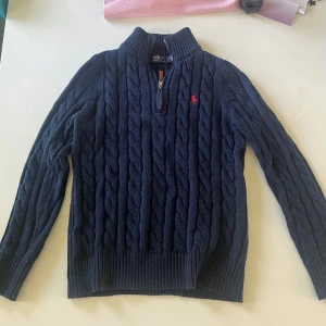 Marinblå kabelstickad tröja Ralph Lauren - Kabelstickad marinblå tröja från Polo Ralph Lauren med halv dragkedja och hög krage. Tröjan har lång ärm, ribbade muddar och röd broderad logga på bröstet. Perfekt för dig som gillar klassisk och stilren look. Pris kan diskuteras. Skriv för fler bilder 