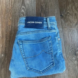 Jacob Cohen Nick Slim blå jeans - Snygga ljusblå jeans från Jacob Cohen i modellen Nick Slim. Jeansen har klassisk femficksdesign, silverfärgade knappar och coola broderade detaljer. Tillverkade i mjuk bomullsdenim med unika insydda mönster på insidan av fickan. Perfekta för dig som gillar stilrena men ändå unika jeans.