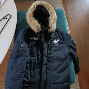 Svart parkajacka från Vinson Polo Club - Svart parkajacka från Vinson Polo Club med stor huva kantad av fejkpäls. Jackan har flera fickor, broderad logga på bröstet och ärmarna samt dragkedja framtill. Perfekt för kalla dagar när du vill hålla stilen och värmen.