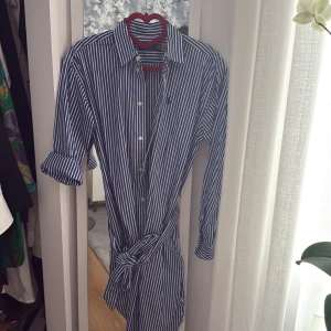 Randig skjortklänning Polo Ralph Lauren - Snygg blå- och vitrandig skjortklänning från Polo Ralph Lauren. Klassisk krage, knappar framtill och avtagbart bälte i midjan. Långärmad modell i mjuk bomull, perfekt för en stilren look.