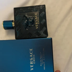 Versace Eros Eau de Toilette 100ml - Versace Eros Eau de Toilette 100ml med ikonisk turkosblå glasflaska och Medusa-detalj i relief. Flaskan har guldiga detaljer och geometriskt mönster. Kommer med matchande blå kartong med guldtext. En klassisk och lyxig herrdoft.