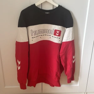 Röd, svart och vit Hummel sweatshirt - Säljer en oversized sweatshirt från Hummel i rött, svart och vitt med stora loggan framtill och klassiska pilar på ärmarna. Tröjan har breda blockfärger, rund hals och långa ärmar. Perfekt för dig som gillar sportig och avslappnad stil.