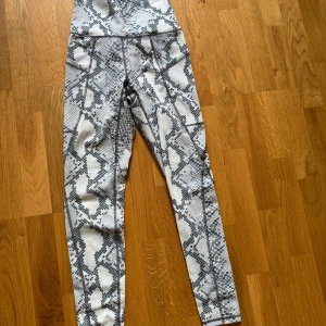 Gråvita tränings tights med ormmönster - Snygga tränings tights  med gråvitt ormmönster och hög midja samt fickor längs benen. Perfekta för dig som vill sticka ut på gymmet eller i vardagen. Stretchigt material som sitter tajt och bekvämt. Passar dig som gillar trendiga och unika träningskläder.