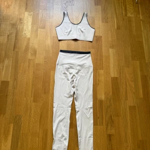 Vit sportig topp med svarta detaljer samt matchande tights till - Säljer en vit sportig topp tillsammans matchande tights med svarta kontrastdetaljer. Toppen har meshpaneler och hakknäppning bak för extra stöd. Perfekt för träning och har en modern, clean look.