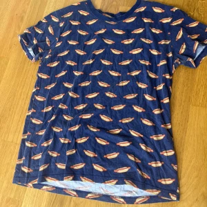 Blå t-shirt med hotdog-mönster XS - Rolig blå t-shirt i storlek XS med ett all-over print av hotdogs. T-shirten har rund halsringning och korta ärmar. Perfekt för dig som gillar unika och lekfulla plagg. Materialet känns mjukt och skönt mot huden.