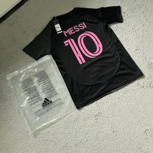 Inter Miami Messi fotbollströja Adidas - Snygg svart Inter Miami fotbollströja från Adidas med rosa detaljer och Messi 10-tryck på ryggen. Tröjan har klubbmärke och Adidas-logga på bröstet, samt korta ärmar. Tillverkad i lätt och ventilerande material, perfekt för match eller träning.