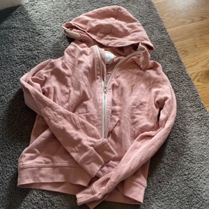 Rosa hoodie med dragkedja från 157 - Mysig ljusrosa hoodie från 157 med huva och hel dragkedja framtill. Tröjan är i mjuk bomull och har långa ärmar samt ribbade muddar. Perfekt för chill dagar eller när du vill ha något bekvämt och snyggt på dig. Dragkedjan kan vara lite trög o hackig!