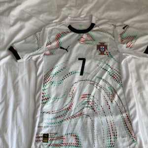 Portugal Ronaldo #7 fotbollströja Puma - Portugal fotbollströja med Ronaldo och nummer 7. Vit bas med gröna och röda prickiga mönster, svart krage och detaljer. Puma-logga på bröstet och officiellt klubbmärke. Tillverkad i lätt och ventilerande material, perfekt för match eller träning. Kan passa på folk som har storlek S också.