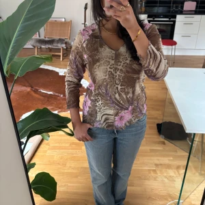 Meshblus med leopard och blommor - Säljer en meshblus i brun leopardmönster med lila och rosa blommor. Blusen har v-ringning, lång ärm och lätt genomskinligt material. Glitter detaljer och vågiga kanter vid ärmslut och nederkant.