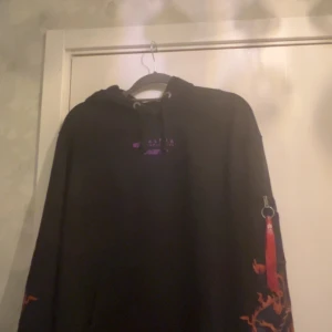 Svart hoodie Alpha Industries med drake - Svart hoodie från Alpha Industries med lila tryck på bröstet och stor färgglad drake i blått, orange och lila på ena ärmen. Snygg röd tag med logga och dragkedja på ärmen. Huva med snörning och klassisk streetwear-vibe. Storlek l passar m