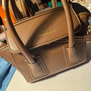 Brun handväska från Fashion & Bags - Snygg brun handväska i strukturerat skinnimitation med guldiga detaljer och dragkedja. Väskan har två rejäla handtag och ett rymligt fack med ljust innerfoder. Perfekt storlek för att få plats med allt du behöver till vardagen.