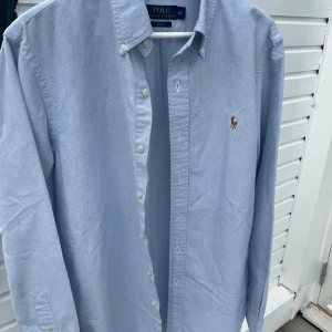 Ljusblå skjorta från Polo Ralph Lauren - Klassisk ljusblå skjorta från Polo Ralph Lauren i 100% bomull. Skjortan har button-down krage, broderad logga på bröstet och vita knappar framtill. Slim fit-modell med lång ärm, perfekt för en clean och stilren look.