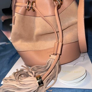 Beige bucket bag från See by Chloé - Säljer en snygg beige bucket bag från See by Chloé med mix av skinn och mocka. Väskan har justerbar axelrem, coola fransdetaljer och guldfärgade metalldetaljer. Perfekt för dig som vill ha en trendig och rymlig väska med lyxig känsla.