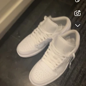 Helvita Nike Jordan 1 sneakers - Säljer ett par helvita Nike Jordan 1 sneakers med klassisk siluett och perforerad tå. Skorna har snörning och är tillverkade i skinn för en clean och fräsch look. Perfekta för dig som gillar stilrena och ikoniska sneakers.