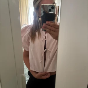 Ljusrosa blus med puffärm och knyt - Supersöt ljusrosa blus med korta puffärmar och två knyt framtill. Blusen är croppad och har en rak passform. Perfekt för dig som gillar romantiska detaljer och en feminin look.