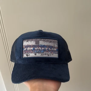 Mörkblå DIF keps  - Snygg mörkblå keps med mesh på baksidan och justerbar snapback. Framtill finns ett stort tryck med bild från läktaren och texten 'FÖR VÅRT LAG' i blått och vitt. Perfekt för dig som gillar sport och vill sticka ut med en unik keps.