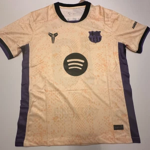 Barcelona Lamine Yamal 10 Nike fotbollströja - Barcelona fotbollströja med Lamine Yamal och nummer 10 på ryggen. Tröjan är beige med ljusorange mönster, svarta och lila detaljer samt svarta tryck. Kortärmad modell i lätt Dri-Fit polyester, med Spotify-logga och UNHCR-tryck. Perfekt för dig som älskar Barca! Pris kan sänkas vid snabb affär!