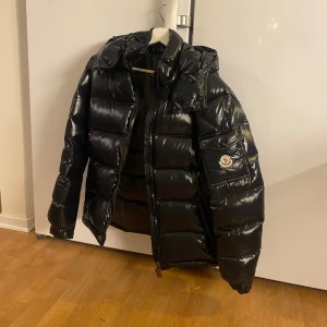Svart Moncler Maya - Säljer en svart pufferjacka från Moncler med glansig finish och huva. Jackan har Moncler-logga på ärmen, dragkedja framtill och quiltad design. Perfekt för kalla dagar och har en cool stil. Fylld med dun och har flera innerdetaljer. Perfekt för Vintern.