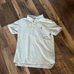Grå pikétröja från Polo Ralph Lauren - Snygg grå pikétröja från Polo Ralph Lauren med klassisk krage och två knappar framtill. Tröjan har korta ärmar och den ikoniska broderade loggan på bröstet. Perfekt för en chill och stilren look. Materialet är mjuk bomull.