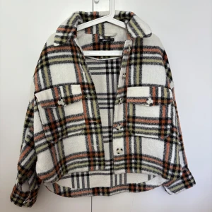 Rutig overshirt från Gina Tricot XS - Säljer en snygg rutig overshirt från Gina Tricot i storlek XS. Jackan har en vit bas med svarta, orange och olivgröna rutor, stora bröstfickor med knappar och klassisk krage. Perfekt att slänga över en hoodie för en chill look.