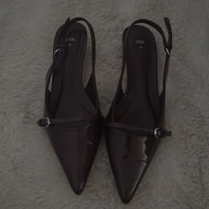 Zara-klackar - Säljer ett par vinröda klackar från Zara i storlek 37. Skorna har en elegant spetsig tå, smal rem med silverfärgat spänne och låg klack. Materialet är glansigt syntet som ger en lyxig vibe. Perfekt för dig som gillar stilrena och trendiga detaljer.