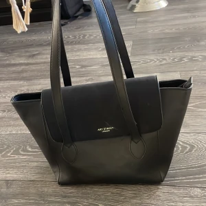 Svart handväska från Art Street London - Snygg svart handväska med stilren design och rejäla handtag. Väskan har ett stort huvudfack, innerfack med dragkedja och en praktisk ficka på baksidan. Tillverkad i skinnimitation och har guldfärgad logga framtill. Perfekt för dig som gillar klassisk och clean look.